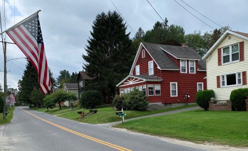 A quiet street lined with houses, featuring a large American flag and green lawns under a cloudy sky.