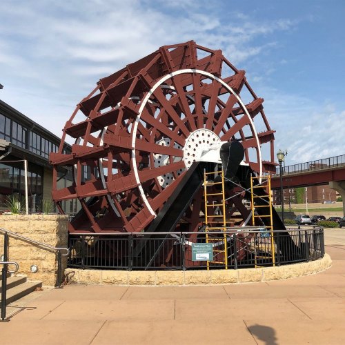 A large, red and white water wheel stands on a stone base, surrounded by a black fence and a paved walkway.