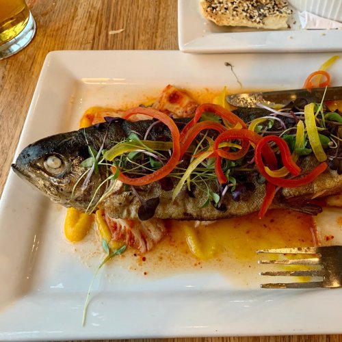 A whole fish garnished with colorful vegetables and microgreens, served on a white plate with a side dish.