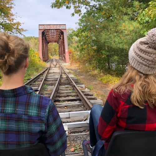 Two people sit on a train track, facing an old bridge surrounded by trees in autumn colors.