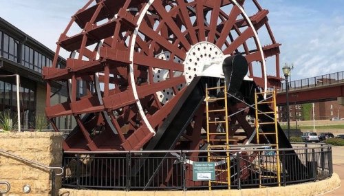 A large, red water wheel structure with a black center, surrounded by a railing and scaffolding, against a blue sky.