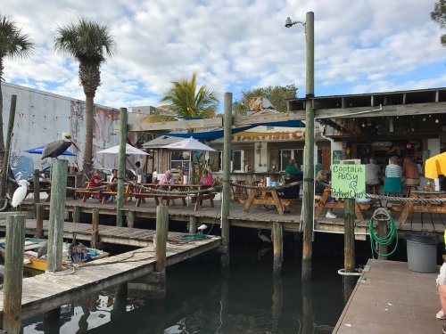A waterfront restaurant with outdoor seating, palm trees, and a dock, bustling with patrons enjoying the view.