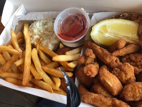 A takeout box filled with fried chicken, French fries, coleslaw, a lemon wedge, and a small cup of dipping sauce.