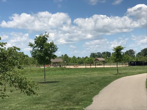 A park scene featuring a path, green grass, trees, and a fenced area with pavilions under a partly cloudy sky.