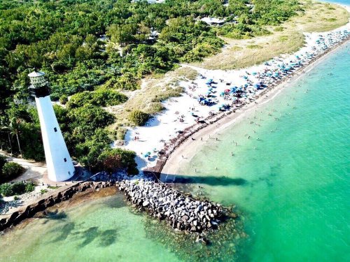 Aerial view of a beach with a lighthouse, surrounded by green trees and colorful umbrellas along the shore.