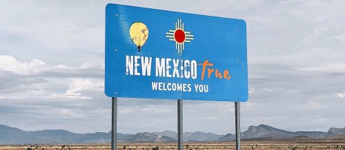 A blue welcome sign for New Mexico featuring a hot air balloon and the state flag, set against a cloudy sky.