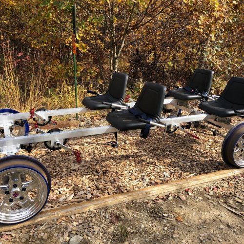 A silver trailer frame with four black seats and large blue wheels, set against a backdrop of autumn foliage.