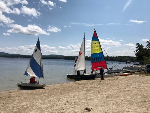 Sailboats on a sandy beach by a calm lake, with people preparing to sail under a clear blue sky.