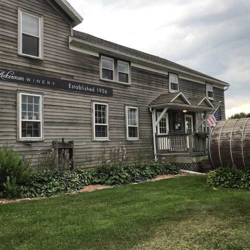 A rustic winery building with a sign reading "Established 1956," surrounded by greenery and a large wooden barrel.