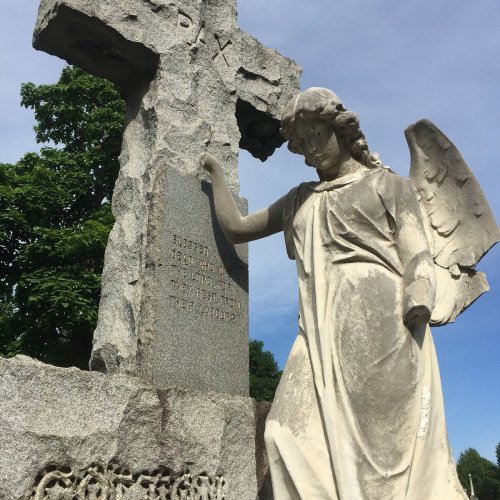 A stone angel leans against a weathered cross, surrounded by greenery under a clear blue sky.
