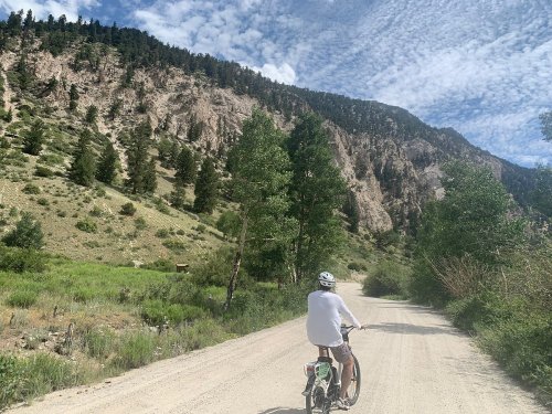 A person rides a bicycle on a dirt road surrounded by green trees and rocky hills under a cloudy sky.