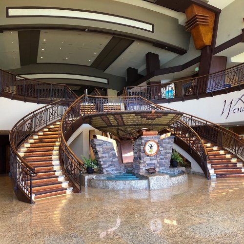 A spacious lobby with a grand staircase, stone features, and elegant wooden accents. Natural light fills the area.