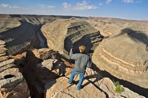 A child stands on a rocky ledge, photographing a vast canyon landscape under a blue sky.