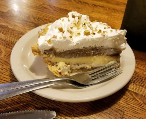 A slice of cream pie topped with whipped cream and nuts on a plate, with a fork beside it on a wooden table.