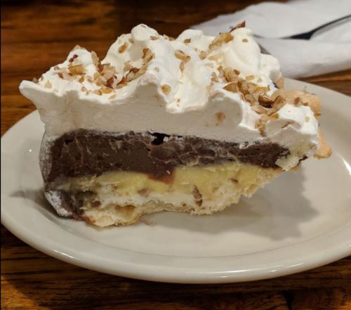 A slice of layered pie with chocolate and banana filling, topped with whipped cream and chopped nuts on a plate.
