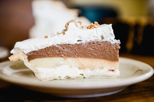 A slice of layered pie with chocolate, vanilla, and whipped cream, topped with nuts on a white plate.