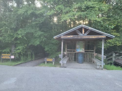 A wooden shelter with benches and a sign, surrounded by trees, leading to a nature trail entrance.