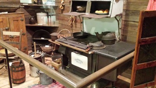 A vintage kitchen scene featuring an old stove, cookware, and various kitchen utensils displayed on wooden shelves.