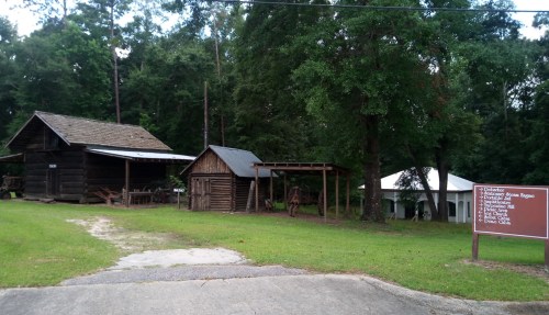 Historic wooden buildings and a tent in a grassy area, surrounded by trees, with a sign listing nearby attractions.