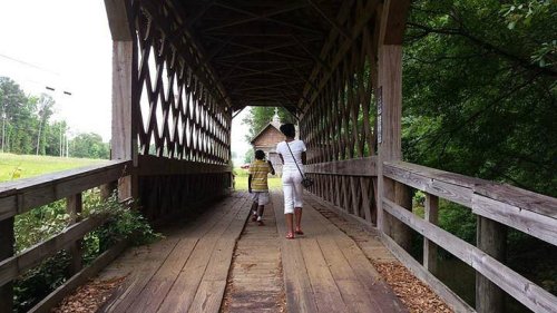 Two people walk through a wooden covered bridge surrounded by greenery.