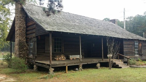 A rustic wooden cabin with a stone foundation, porch, and stacked firewood, surrounded by greenery.