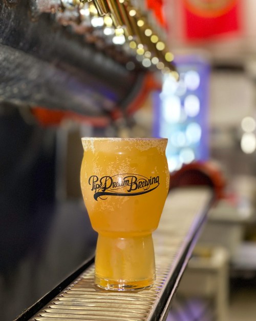 A frosty glass of beer sits on a bar, featuring the logo of Pipe Dream Brewing in a vibrant setting.