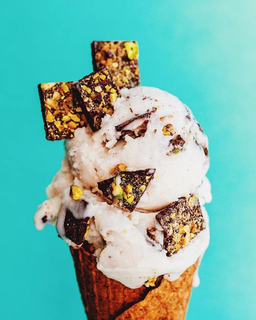 A close-up of a cone filled with creamy ice cream topped with chocolate pieces and nuts against a bright turquoise background.