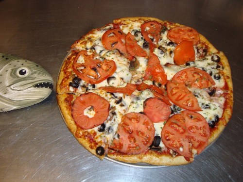 A pizza topped with tomatoes, olives, and cheese, next to a ceramic fish decoration on a metal surface.