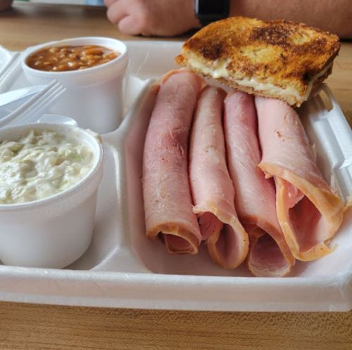 A takeout tray with sliced ham, a toasted sandwich, baked beans, and coleslaw in separate containers.