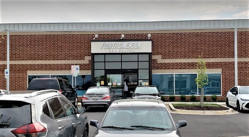 Exterior view of a brick building with large windows, featuring a sign that reads "Fourth Dimension" and parked cars in front.