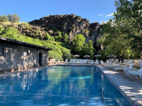 A serene pool surrounded by lush greenery and rocky hills under a clear blue sky. Sun loungers line the poolside.