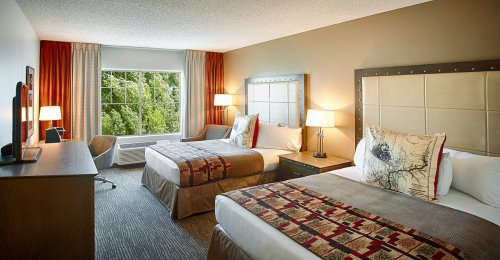 A cozy hotel room with two beds, a desk, and a large window letting in natural light. Decorated in warm tones.