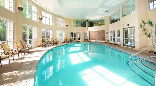 Indoor swimming pool with large windows, lounge chairs, and bright sunlight reflecting on the water.