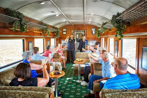 A group of passengers toasting with drinks inside a vintage train car, surrounded by scenic views.