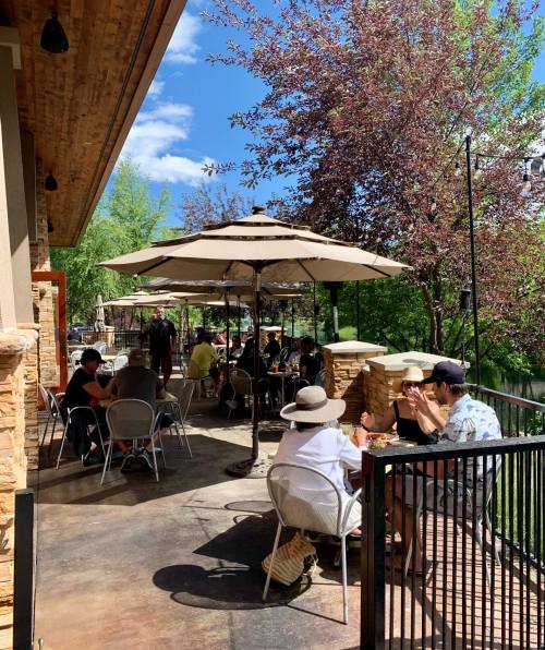 Outdoor dining area with patrons under umbrellas, surrounded by greenery and a sunny sky.