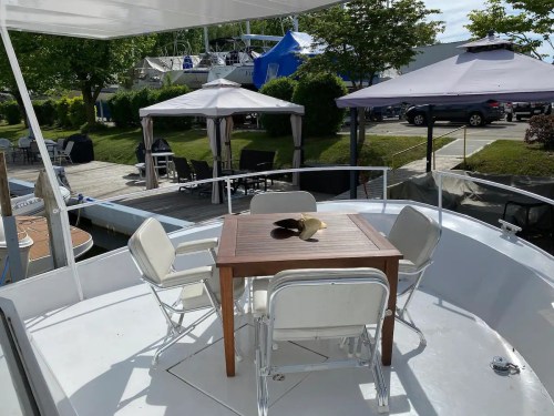 A boat deck with a wooden table and chairs, overlooking a marina with boats and green trees in the background.