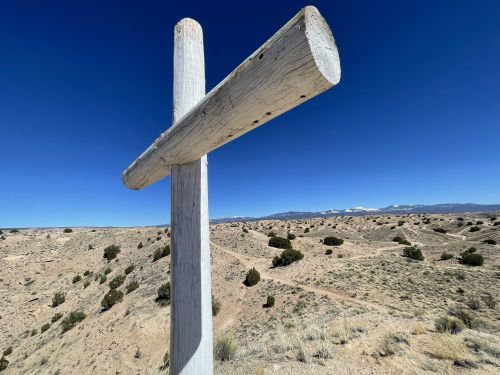 A large wooden cross stands on a hillside, with a clear blue sky and distant mountains in the background.