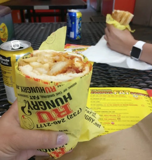 A hand holding a wrapped burrito filled with fries and chicken, with drinks and a menu in the background.