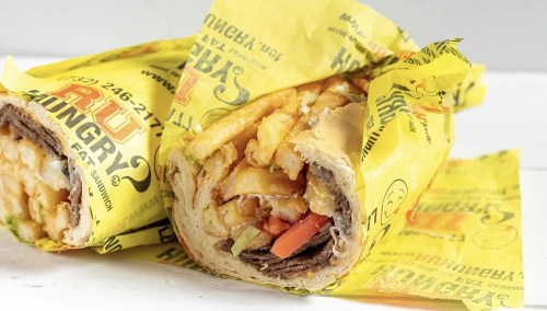 A wrapped sandwich filled with meat, fries, and vegetables, resting on yellow branded paper.