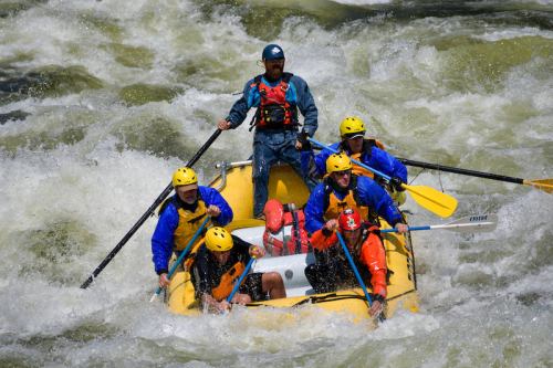 A group of six people in yellow helmets navigates a white-water raft through turbulent rapids.