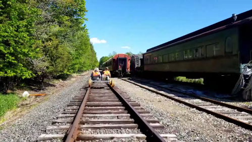 Two people on a small rail cart traveling along a train track beside vintage train cars and lush greenery.