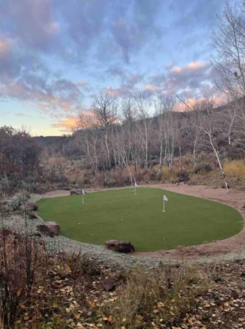 A small, green golf putting green surrounded by trees and rocky terrain under a cloudy sky.