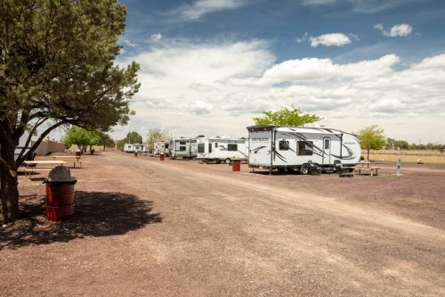 A row of RVs parked along a gravel road, with trees and a picnic area under a partly cloudy sky.