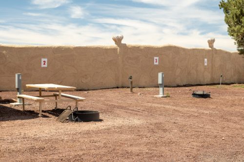 A gravel area with a picnic table, fire pit, and numbered posts, surrounded by a low wall under a blue sky.