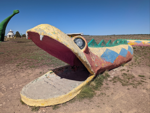 A colorful, large snake sculpture with an open mouth, set against a clear blue sky and grassy landscape.