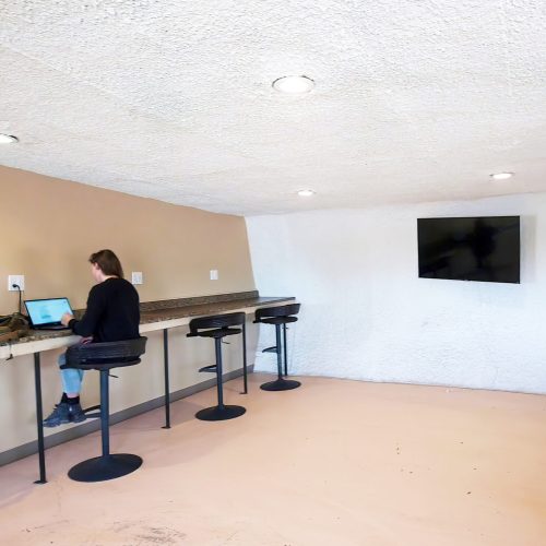 A person sits at a counter with a laptop in a minimalist room featuring a TV and bar stools.
