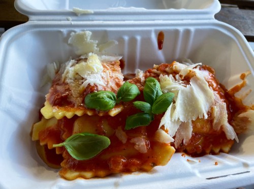 A takeout container with ravioli topped with marinara sauce, cheese, and fresh basil leaves.