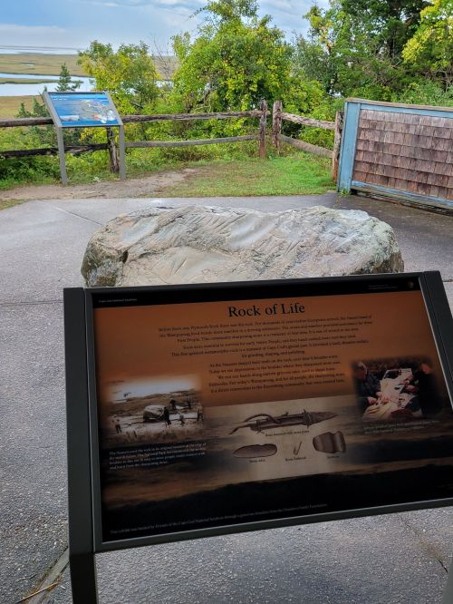 A large rock labeled "Rock of Life" near an informational sign, surrounded by greenery and a scenic view.