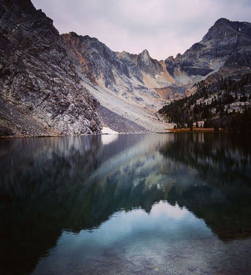 A serene mountain lake reflecting rugged peaks and a cloudy sky, surrounded by rocky terrain and sparse trees.