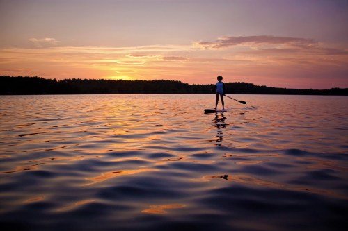A person paddleboarding on a calm lake at sunset, with vibrant colors reflecting on the water.
